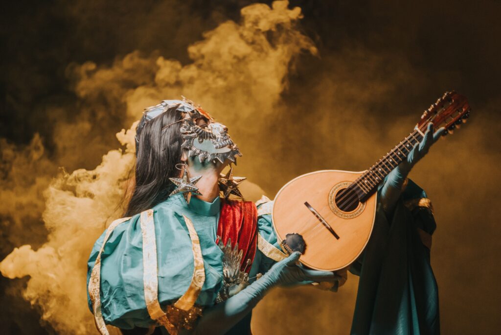Masked performer in colorful costume playing mandolin amid dramatic smoke.