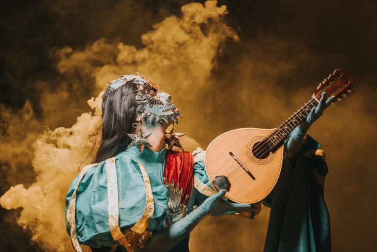Masked performer in colorful costume playing mandolin amid dramatic smoke.